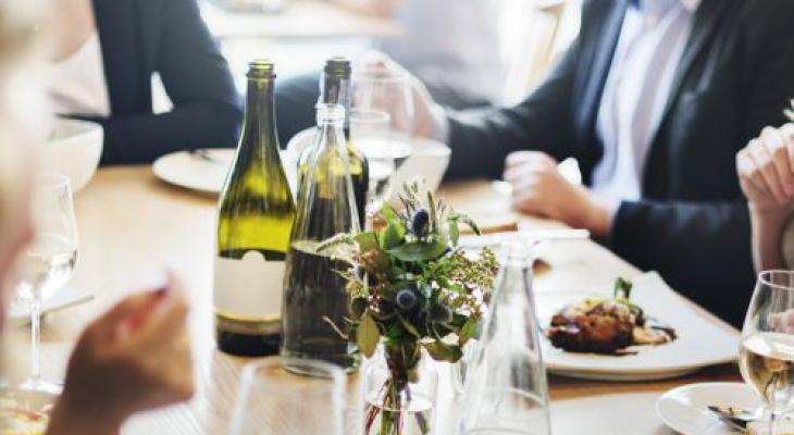 Persone a tavola con bottiglie di vino e piatti di cibo, atmosfera conviviale.