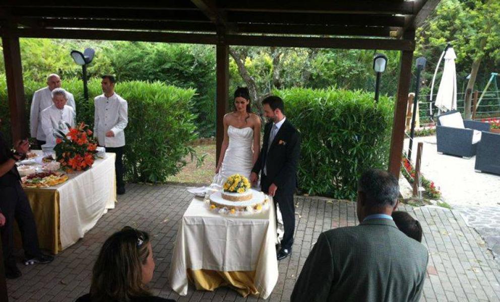 Sposi tagliano la torta nuziale all'aperto, circondati da ospiti e personale di servizio.
